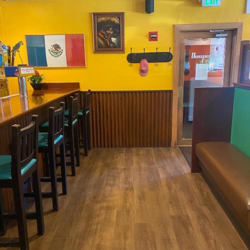 Interior of a colorful restaurant with a bar, wooden seating, and a portrait on the wall. Bright yellow walls.