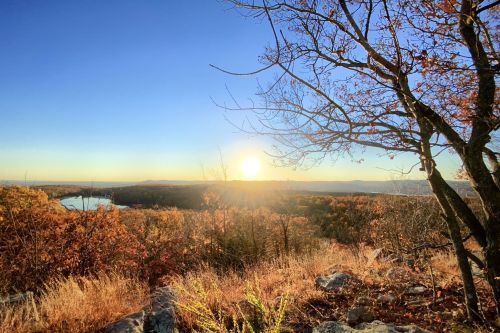 Sunrise over a scenic landscape with autumn foliage, trees, and a calm body of water in the distance.
