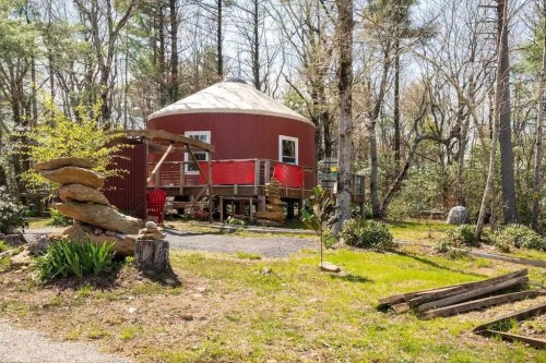 A red yurt surrounded by trees, with a wooden deck and stone sculptures in a grassy area.