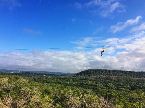 A person zip-lining over lush green hills under a blue sky with scattered clouds.