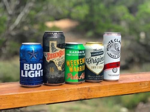 Five different beer cans lined up on a wooden surface, surrounded by greenery.