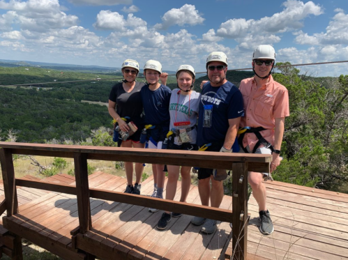 A group of five people in helmets and harnesses stand on a wooden platform with a scenic view of green hills and blue sky.