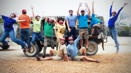 A group of people in colorful shirts and helmets joyfully posing and jumping in front of a truck in an outdoor setting.