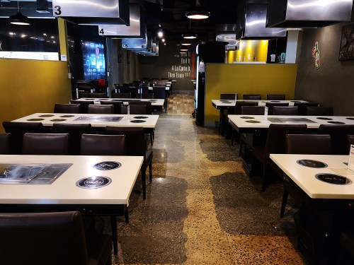 Interior of a modern restaurant with tables featuring built-in grills, dark seating, and a yellow accent wall.