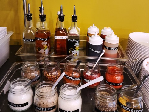 A variety of sauces and spices in jars and bottles, including vinegar, soy sauce, and chili powder, on a countertop.