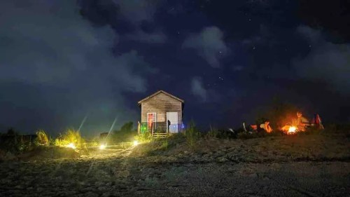 A small wooden house at night, surrounded by clouds and stars, with lights illuminating the path and a campfire nearby.