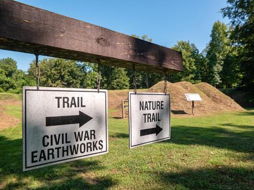 Two signs hanging from a wooden post indicate directions for "Civil War Earthworks" and "Nature Trail" in a green park.