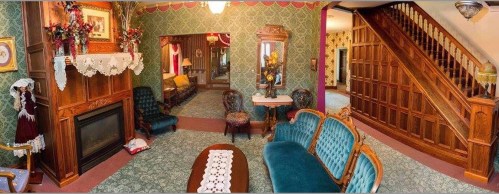 A vintage living room with ornate furniture, a fireplace, and a staircase, featuring patterned wallpaper and decorative accents.