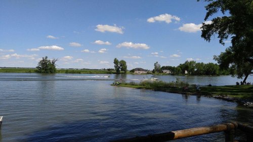 A serene river scene with boats, lush greenery, and a clear blue sky dotted with clouds.