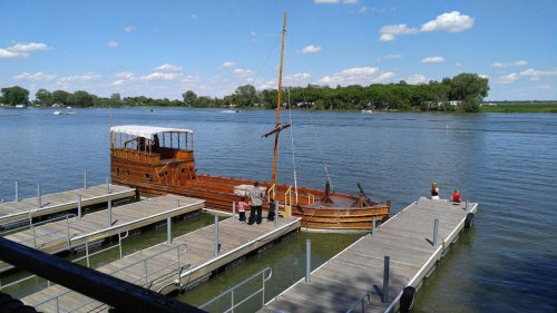 A wooden boat docked at a pier on a calm river, with people enjoying the sunny day and blue sky.