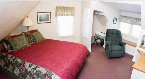 Cozy bedroom with a red floral bedspread, a green armchair, and natural light from two windows.