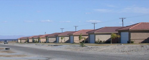 Row of single-story houses with red tile roofs along a quiet street under a clear blue sky.