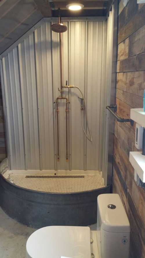 A rustic bathroom featuring a metal shower stall, a showerhead, and a toilet against a wooden wall.