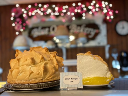 A whole lemon meringue pie and a slice, displayed in a bakery with decorative lights in the background.