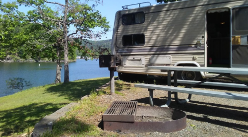 A parked RV by a serene lake, with a picnic table and a fire pit nearby, surrounded by trees.