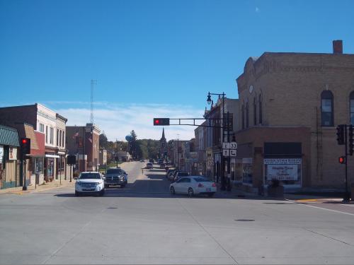 A small town street with shops, traffic lights, and clear blue skies. Cars are stopped at a red light.