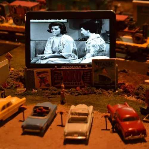 A miniature scene featuring toy cars and a vintage TV displaying a black-and-white show with two women.