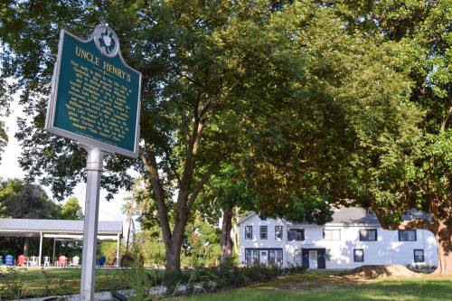 A historical marker for Uncle Henry's stands in front of a large tree and a white building in a green landscape.