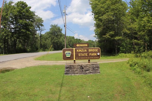 Sign for Kinzua Bridge State Park, surrounded by greenery, with a road and power lines in the background.