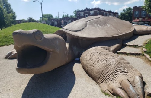 A large, sculpted turtle lies on a grassy area, with its mouth open and details of its shell and limbs visible.