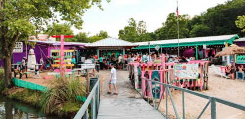 A vibrant outdoor dining area by a lake, featuring colorful buildings, a wooden walkway, and people enjoying the atmosphere.