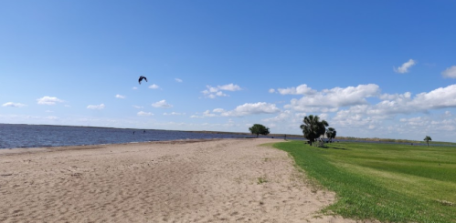 A sandy beach by a calm lake, with green grass and trees under a blue sky with scattered clouds.