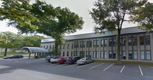 A modern building with large windows and a covered entrance, surrounded by trees and parked cars in the foreground.