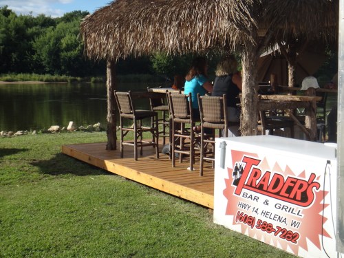 A thatched-roof bar and grill by a river, with patrons seated at high chairs on a wooden deck.
