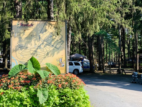 A campground map sign surrounded by greenery and flowers, with tents and vehicles visible in the background.