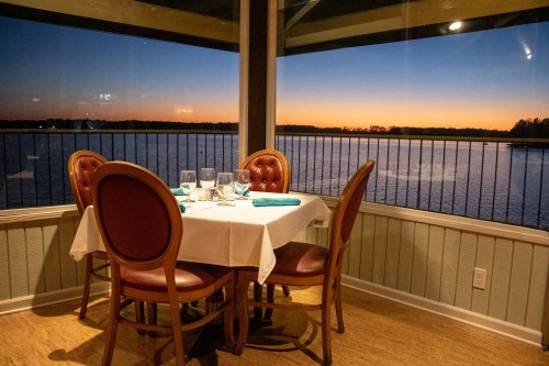 A cozy dining table set for four, with a view of a serene lake at sunset through large windows.