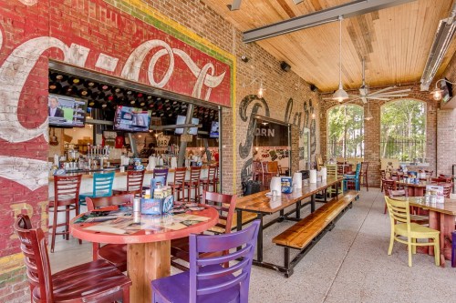 Outdoor dining area of a restaurant with colorful chairs, exposed brick walls, and a bar in the background.
