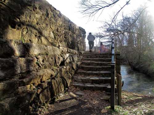 A person walks up stone steps beside a creek, with a stone wall and trees in the background.