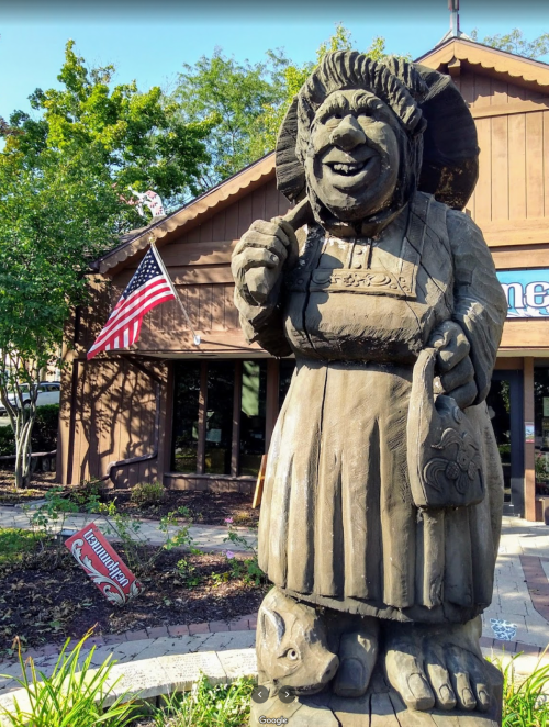 A large wooden statue of a smiling woman in a dress, holding a bag, stands outside a building with an American flag.