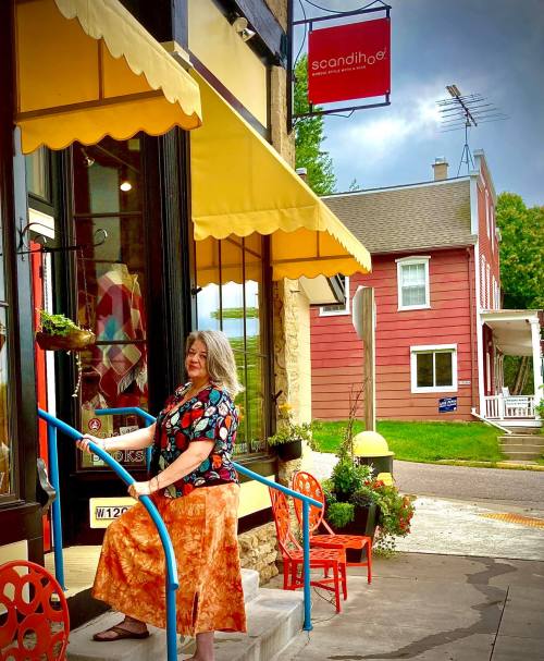 A woman with long hair sits on steps outside a colorful shop, wearing a patterned top and orange skirt, with chairs nearby.