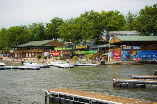 A lakeside view featuring a restaurant named "Dog Days" and several boats docked at wooden piers. Green trees in the background.