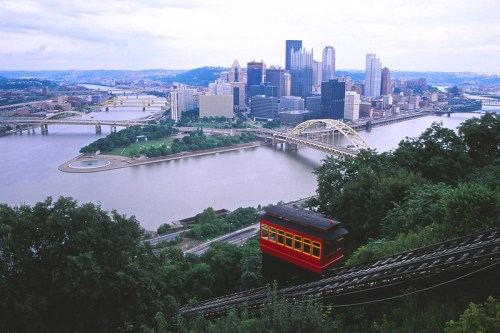 A scenic view of Pittsburgh with a funicular railway, bridges, and skyscrapers along the river.