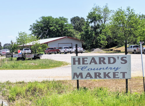 Sign for Heard's Country Market with a gravel parking lot and several vehicles in the background, surrounded by greenery.