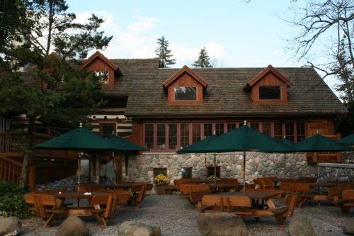 A rustic log cabin with a stone foundation, surrounded by trees and outdoor seating under green umbrellas.
