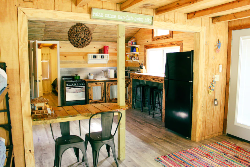 Cozy wooden interior with a kitchen, dining area, and a sign that says "relax, canoe, nap, fish, swim, hike."