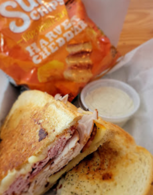 A close-up of a grilled sandwich with turkey and cheese, accompanied by a bag of chips and a small container of dip.
