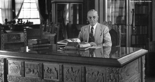 A man in a suit sits at an ornate wooden desk, surrounded by books in a library setting.
