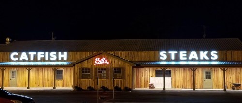 A wooden restaurant exterior at night with illuminated signs reading "CATFISH" and "STEAKS."