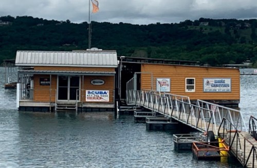A floating building with a wooden deck on a lake, featuring signs for scuba diving and other activities.