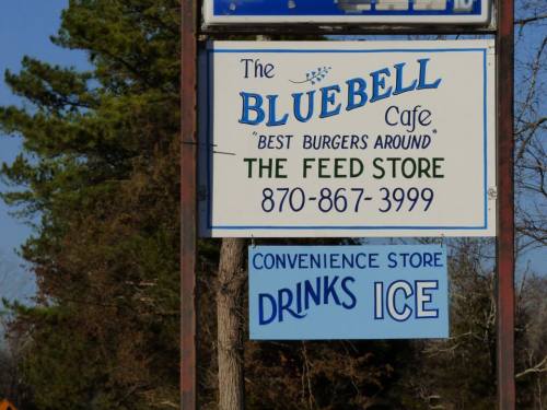 Sign for Bluebell Cafe featuring "Best Burgers Around," along with The Feed Store and a convenience store offering drinks and ice.