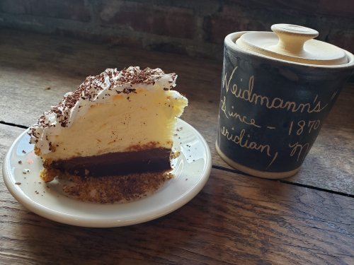 A slice of layered dessert with whipped cream and chocolate, next to a decorative container labeled "Weidmann's 1870."