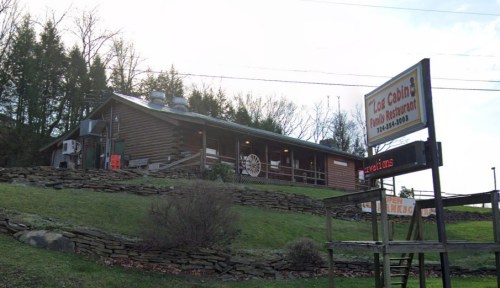 A rustic log cabin restaurant with a sign, surrounded by greenery and trees under a partly cloudy sky.