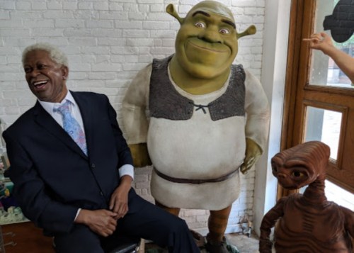 A man in a suit sits next to Shrek and E.T., smiling in a bright room with a window.