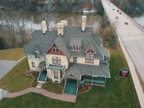 Aerial view of a large, historic house with multiple chimneys, surrounded by greenery and near a river and road.