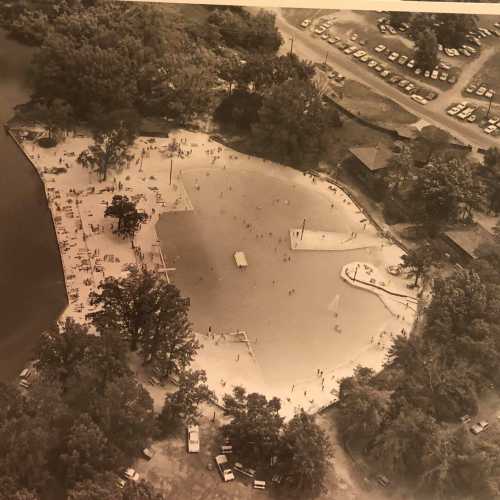 Aerial view of a beach area with people swimming, surrounded by trees and parking spaces in the background.