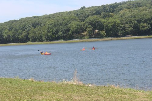 Three kayakers paddle on a calm lake surrounded by green hills and trees under a clear blue sky.